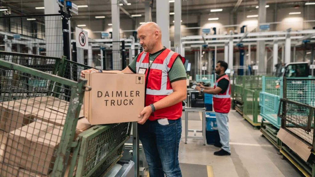 A warehouse employee in a high-visibility red vest processes a box labeled "Daimler Truck" inside the automated Halberstadt Global Parts Center. (Photo: Daimler Truck)