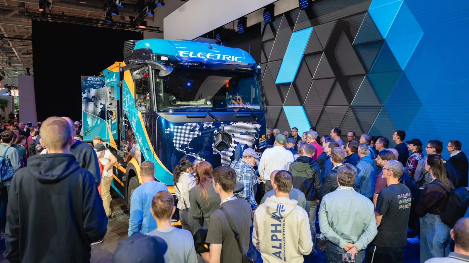 A large crowd of trade attendees standing in front of a blue Mercedes-Benz electric heavy truck on an exhibition floor.