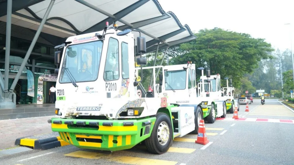 A row of white and green Terberg electric terminal tractors parked single-file along the driveway outside a modern port building.
