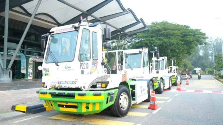A row of white and green Terberg electric terminal tractors parked single-file along the driveway outside a modern port building.
