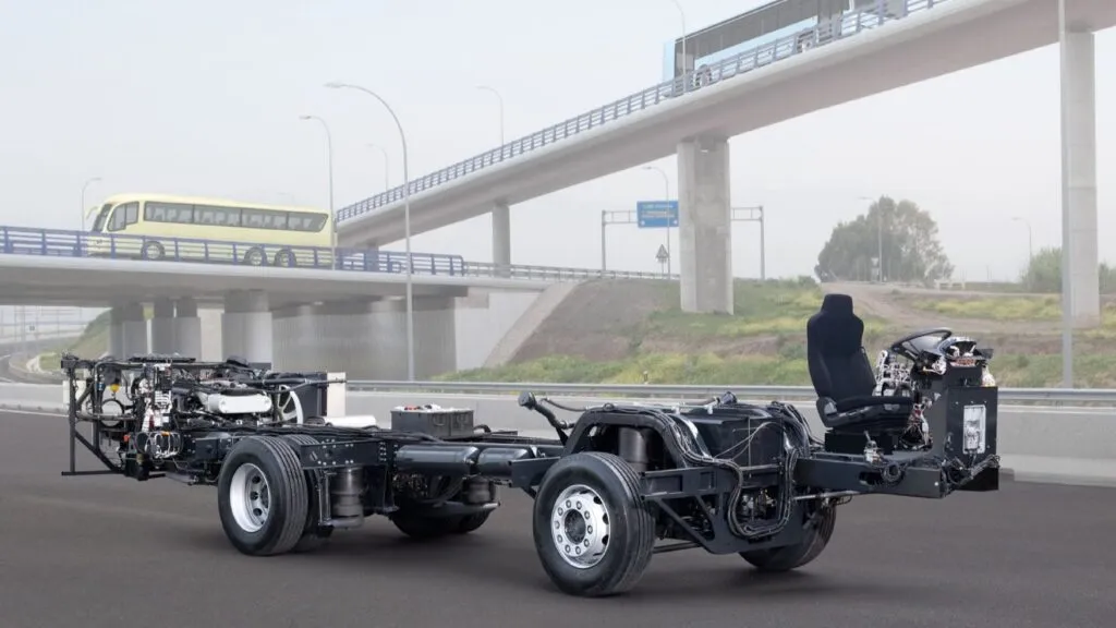 A bare MAN coach bus chassis showing the driver's seat, steering assembly, engine components, and wheels parked on an asphalt road in front of an overpass.