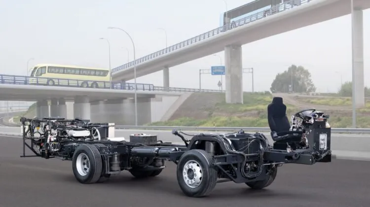 A bare MAN coach bus chassis showing the driver's seat, steering assembly, engine components, and wheels parked on an asphalt road in front of an overpass.
