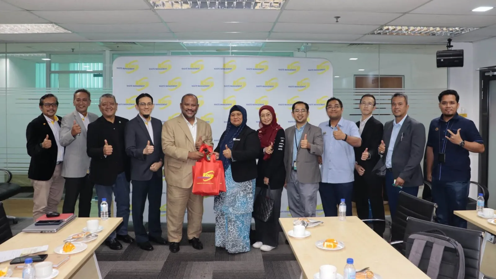 A group of corporate and government representatives standing together giving thumbs-up gestures inside a meeting room, with two leaders holding a gift bag in the centre.