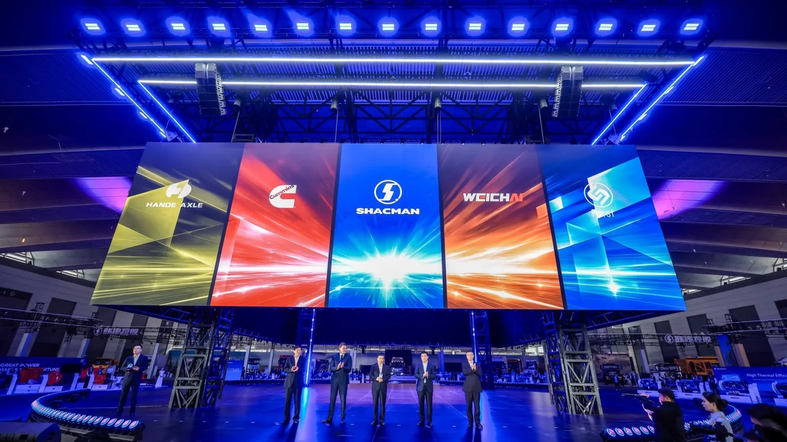 Five executives in business suits standing on a stage illuminated by blue lights in front of a giant digital screen displaying the logos of HanDe Axle, Cummins, Shacman, Weichai, and Fast.