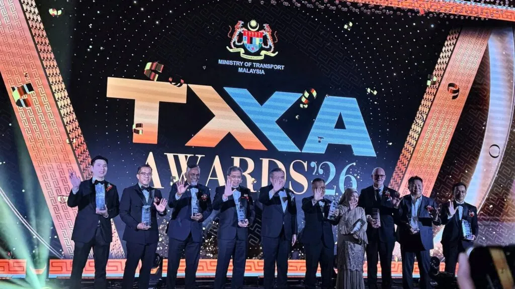 Swift Group CEO Loo Yong Hui and other corporate leaders in black tuxedos holding trophies and waving on stage under the Ministry of Transport Malaysia TXA Awards '26 digital screen.