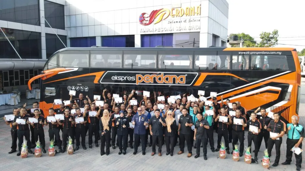 A group photo of express bus drivers in uniform holding completion certificates posing alongside JPJ officers and corporate executives in front of a black and orange Ekspres Perdana double-decker bus.