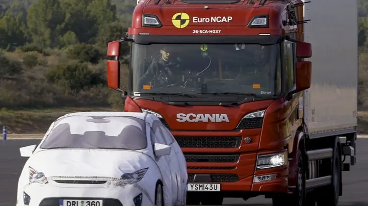 A red Scania P-series truck on a test track undergoing Euro NCAP crash avoidance testing behind a white target vehicle dummy.