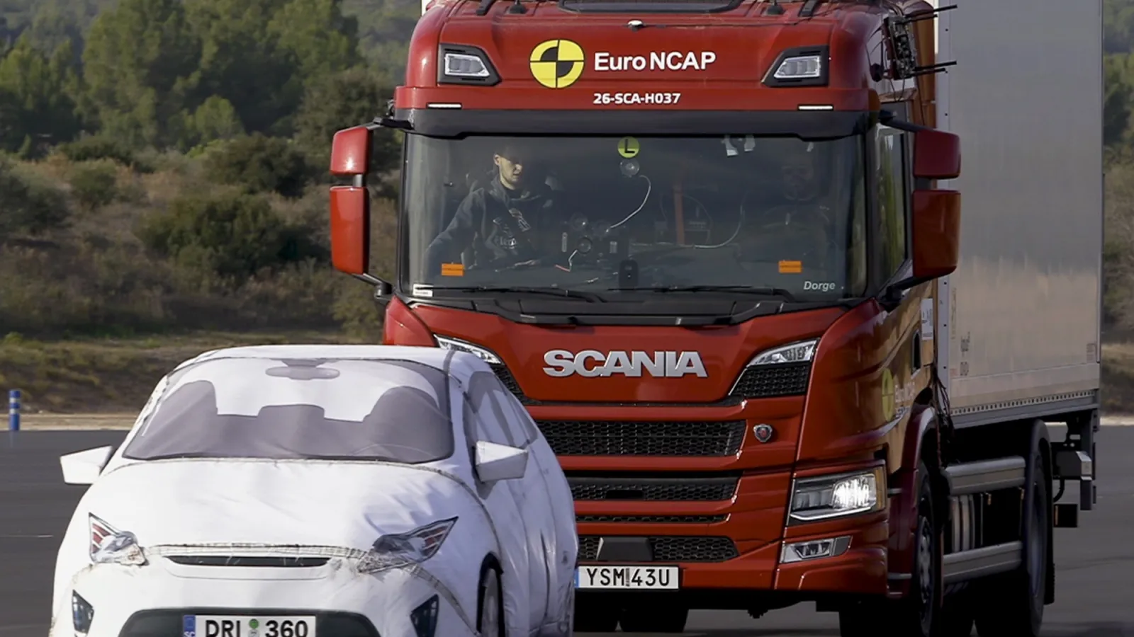 A red Scania P-series truck on a test track undergoing Euro NCAP crash avoidance testing behind a white target vehicle dummy.