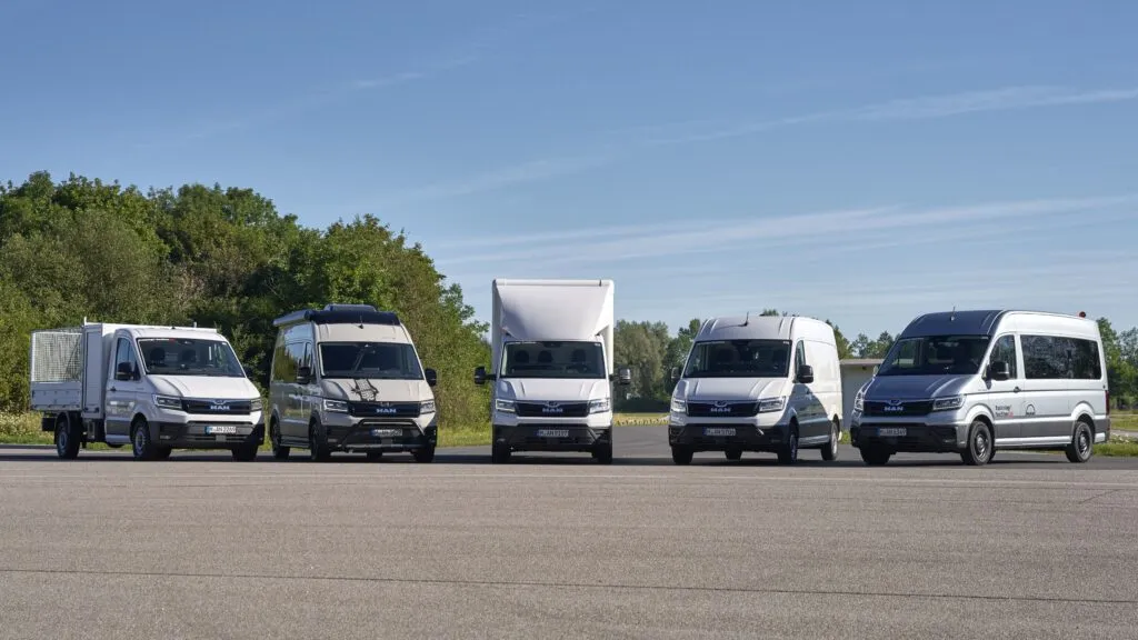 Five white and silver MAN TGE vans in different body configurations lined up on an asphalt test track under a clear blue sky.